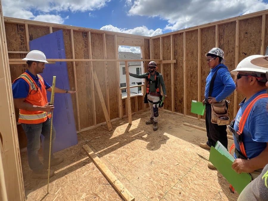 A fall protection class gathered around a framed training structure while an instructor in a full-body harness demonstrates at a wall opening