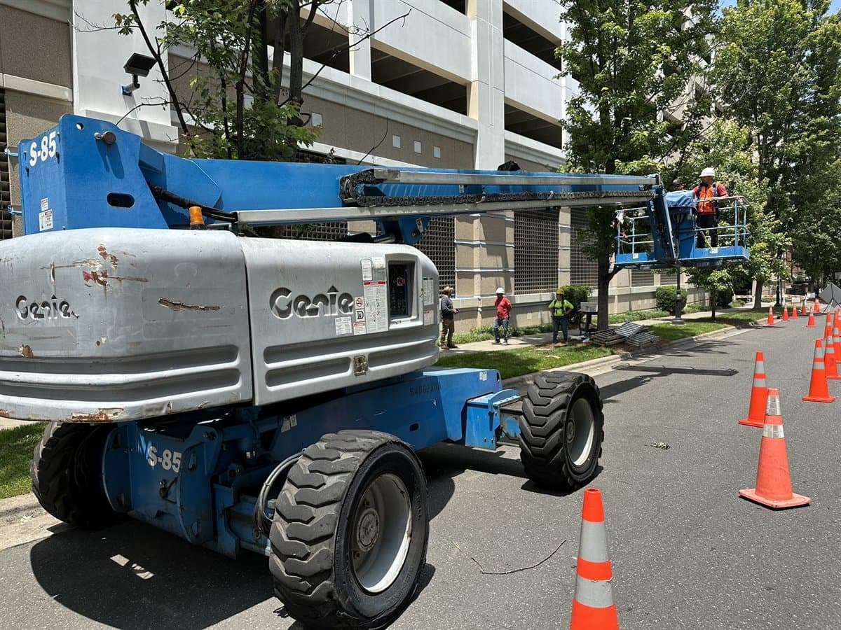 Two workers in hard hats and high-visibility vests working from the platform of a telescopic boom lift raised over a city street, with cones marking off the work zone below