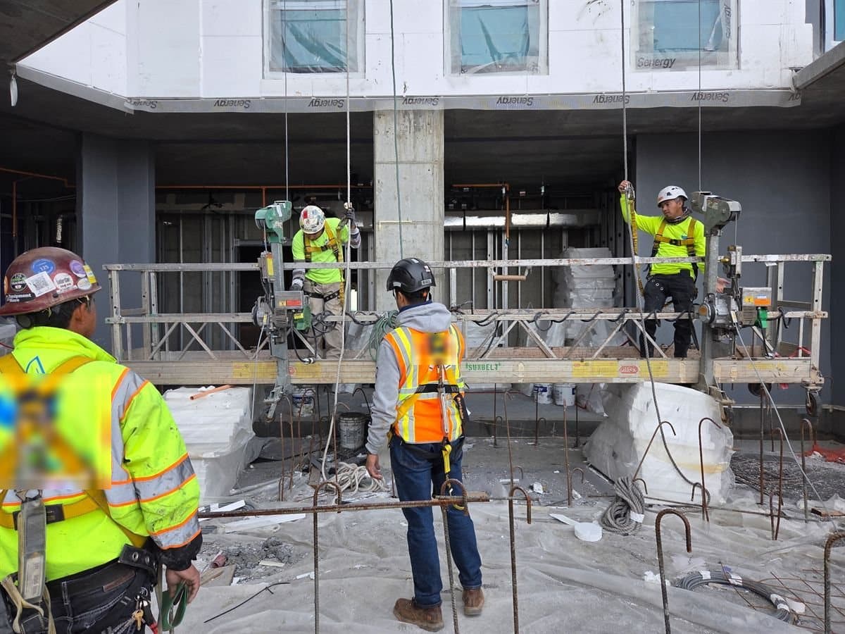 A crew working from a suspended swing stage scaffold, tied off in full-body harnesses, on a commercial building